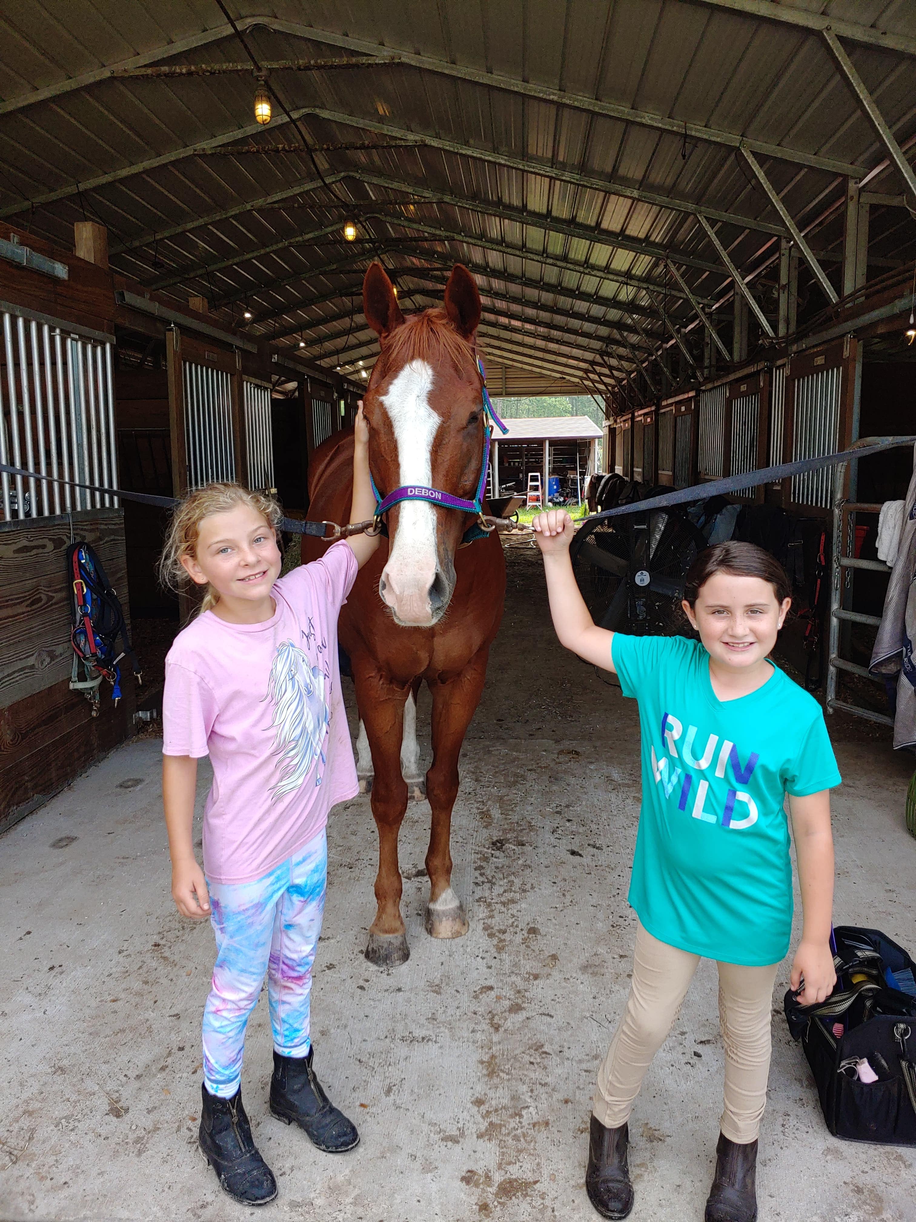 Two summer camp students standing the aisle of the main barn with their favorite horse at the crossties.