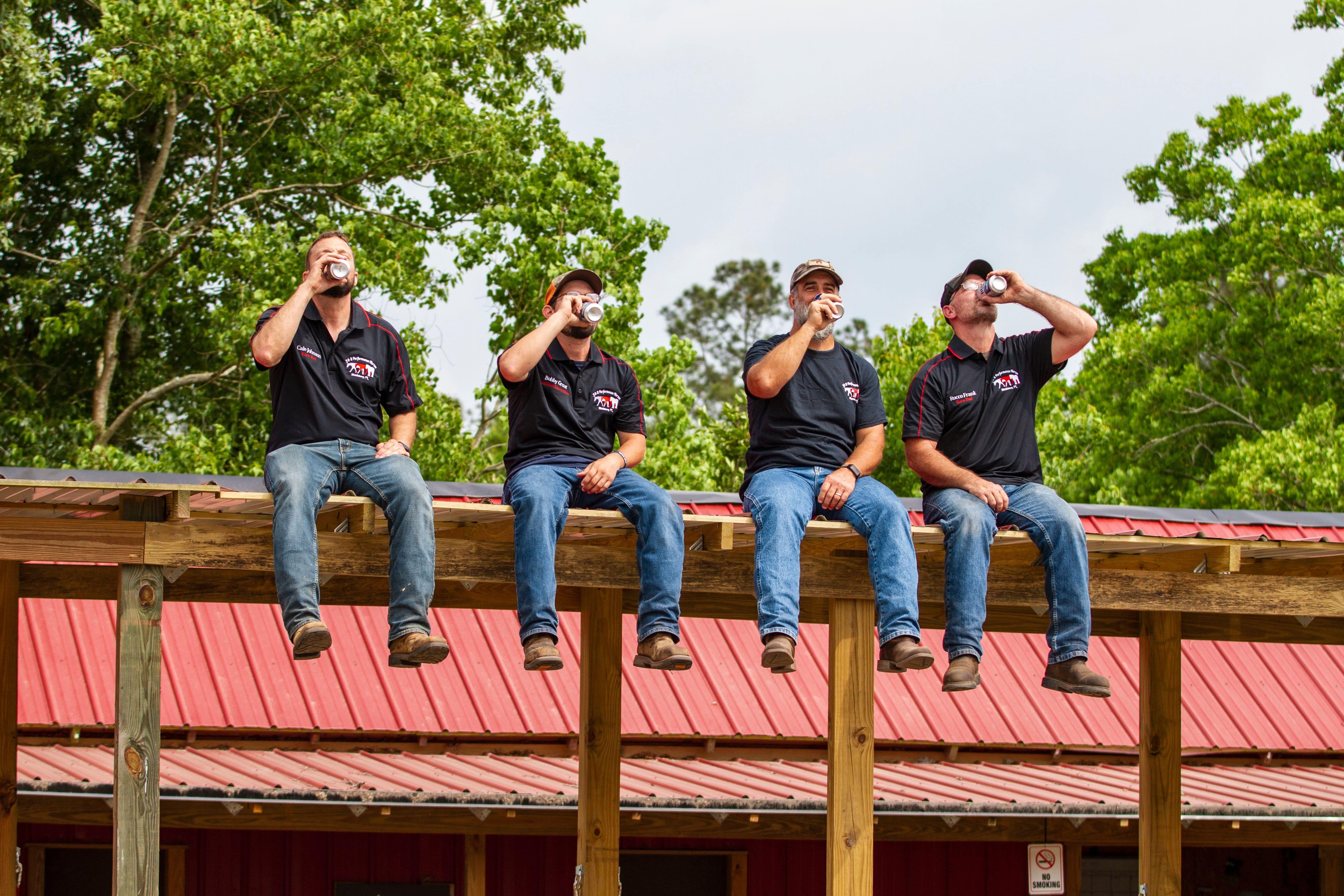 Four barn dads are sitting on the roof of the crossties they built. All four are wearing J & S shirts while drinking out of cans.